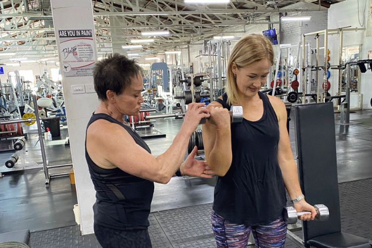 Taylor Young and a woman engaged in a training session at Supershape Gym, highlighting the supportive environment of the ladies-only section. One woman demonstrates proper form while lifting a dumbbell, while Taylor Young provides guidance and encouragement.