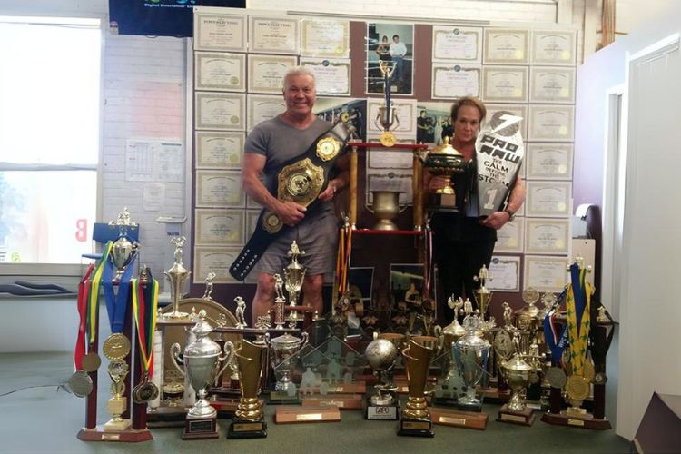 A photo of Laurie and Taylor, standing proudly among numerous fitness trophies and awards at Supershape Gym. Laurie holds championship belts, while Taylor displays a large trophy, showcasing their achievements in the fitness industry. Behind them, certificates and additional trophies highlight their expertise and success.